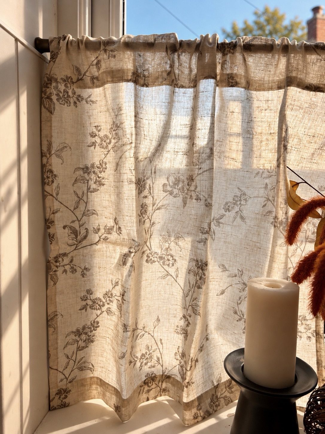 Short beige floral linen curtains hanging over a sunny window with a white candle.