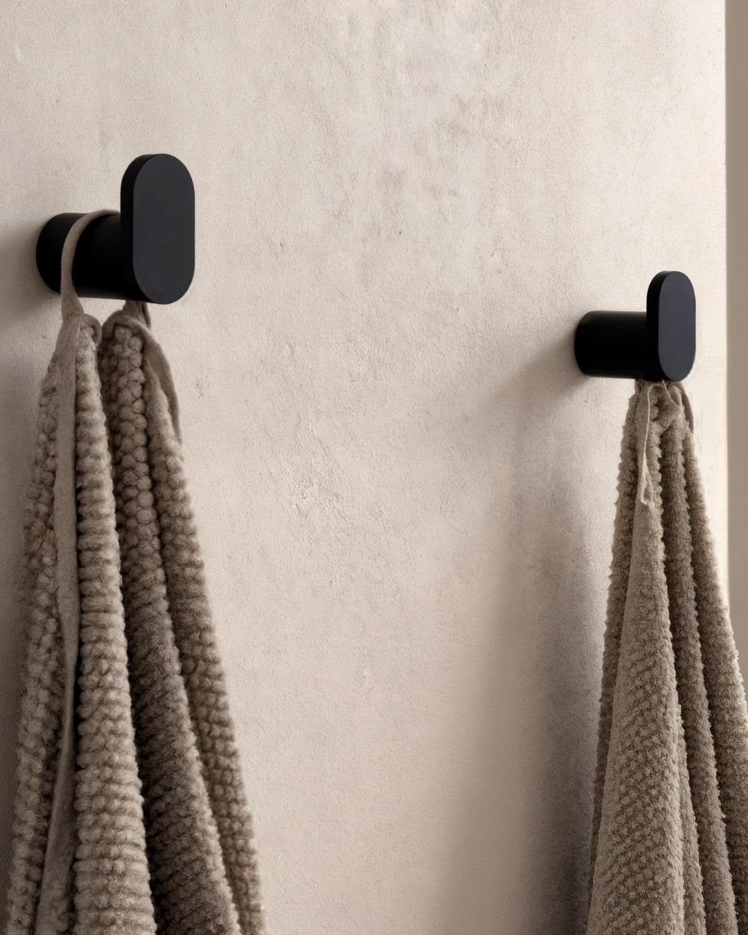 Two matte black towel hooks holding beige towels on a textured wall.